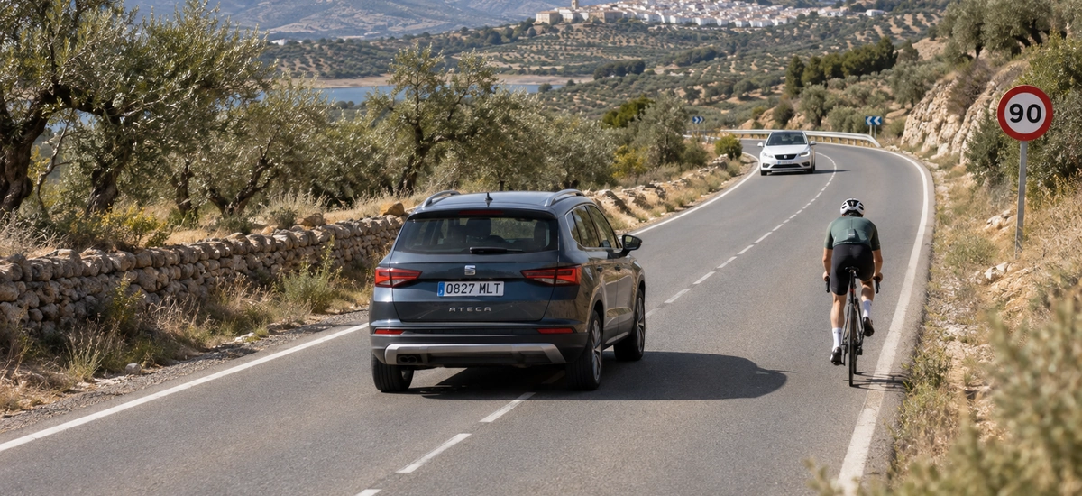 Coche adelantando a un ciclista por el carril contrario en una carretera convencional con señal de 90, mientras se acerca otro vehículo, según las normas del Real Decreto 518/2026
